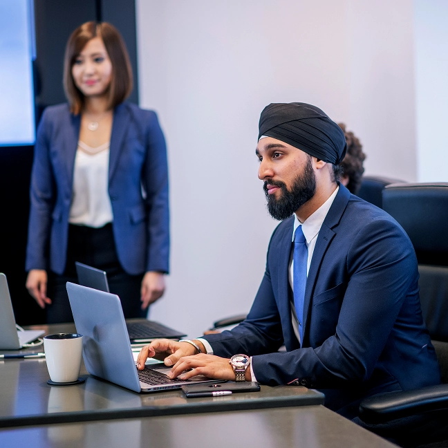 Businessman wearing a black turban working on a laptop at an office desk, with a colleague standing in the background